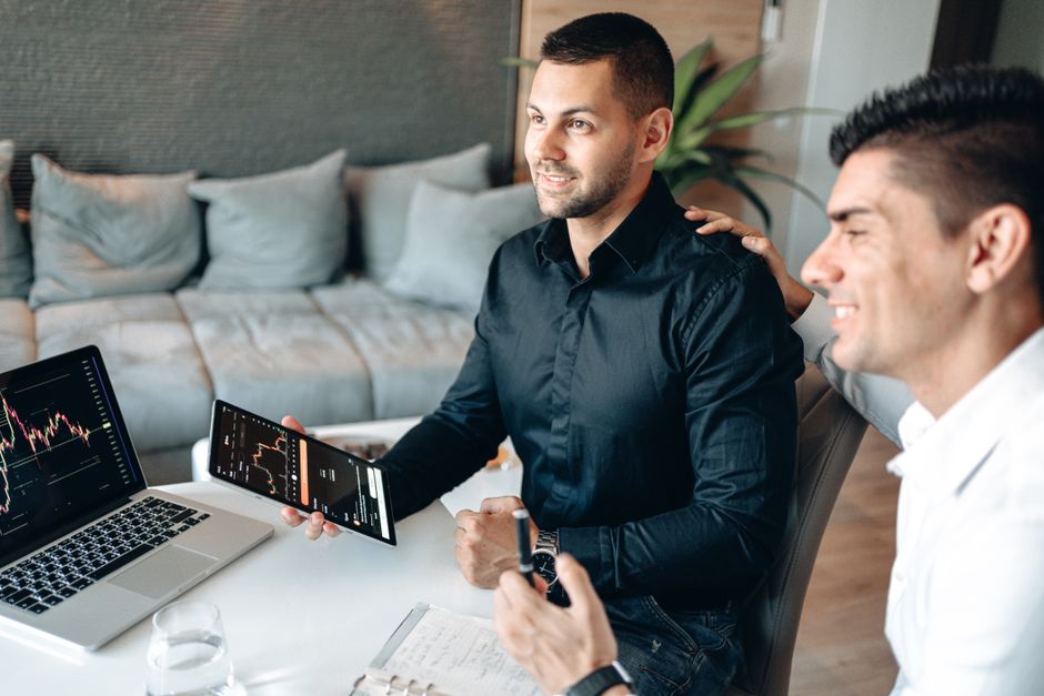A pair of men engaged in a discussion in an office setting, with one man holding a smartphone displaying stock or financial charts, while the other man listens attentively. On the table in front of th