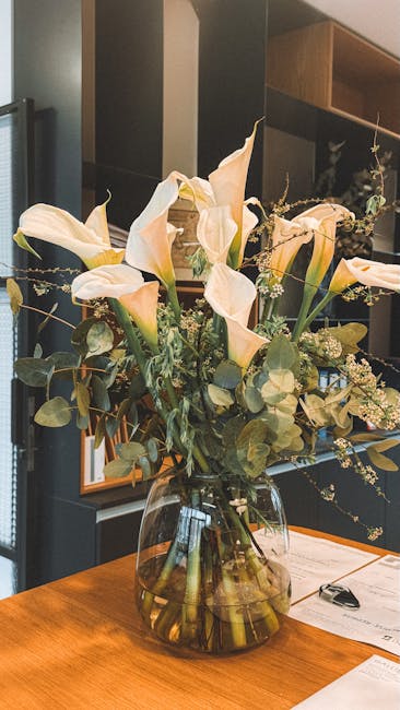 A clear glass vase with a wide base holding a floral arrangement of cream-colored calla lilies with soft, trumpet-shaped petals and dark green stems. The bouquet includes sprigs of small white baby's 