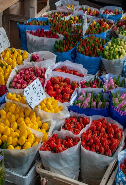 A vibrant display of fresh tulips arranged in large open paper bags at a flower market. The tulips come in a variety of colours including bright yellow, soft pink, rich red, coral, and shades of purpl