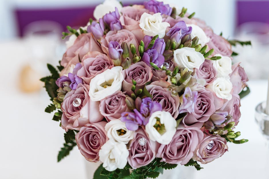 A close-up of a floral bouquet featuring pale pink roses, white lisianthus, and purple freesias, arranged in a compact, round style with green foliage accents. The flowers appear fresh with delicate,