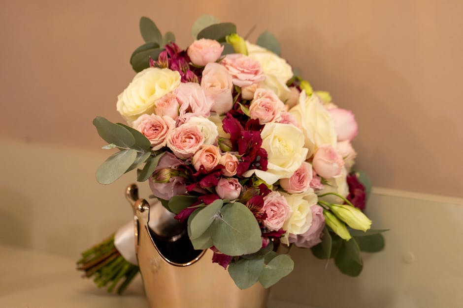 A floral bouquet featuring pale pink roses, white roses, and deep burgundy flowers, accented with soft green eucalyptus leaves, arranged in a rounded, lush style. The bouquet is displayed in a silver