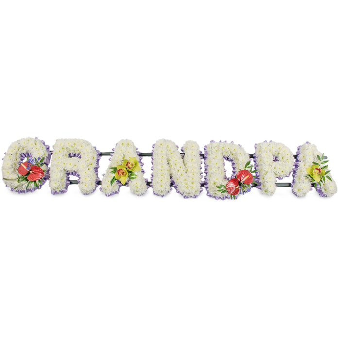 The image shows a long, carefully crafted floral tribute spelling out "GRANDPA" in bold, upright capital letters, displayed against a plain white background so every nuance of the arrangement is easy to see. Each letter is made from countless small white chrysanthemums, packed tightly together to form thick, cushion-like blocks of flowers. Their rounded petals create a soft, almost plush surface, suggesting peace, purity, and gentle remembrance. Along the base of each letter, a narrow border of soft lavender ribbon adds a delicate line of colour; this light purple trim subtly frames the white blooms and gives the design a polished finish without distracting from the word itself. The letters are supported on dark green bars that run horizontally beneath them, visible only in glimpses between the shapes, giving structure while still looking natural. Four focal sprays add colour and personality to the tribute. On the 'G' at the far left and the 'D' in the middle-right, striking clusters of deep red anthuriums stand out, their heart-shaped, glossy petals and central spadices shining against lush green foliage and small touches of purple filler flowers. These rich crimson tones suggest strong, enduring love. The first 'A' and the final 'A' are decorated with clusters of yellow-green orchid blooms, probably cymbidiums, their elegant, open faces and graceful stems tucked into green leaves to create a sophisticated contrast with the white base. The tribute feels bespoke and heartfelt, the sort of personalised "Grandpa" lettering a family in Ealing might place by the coffin or at the graveside to honour a much-loved grandfather, expressing gratitude, respect, and a lifetime of shared stories.